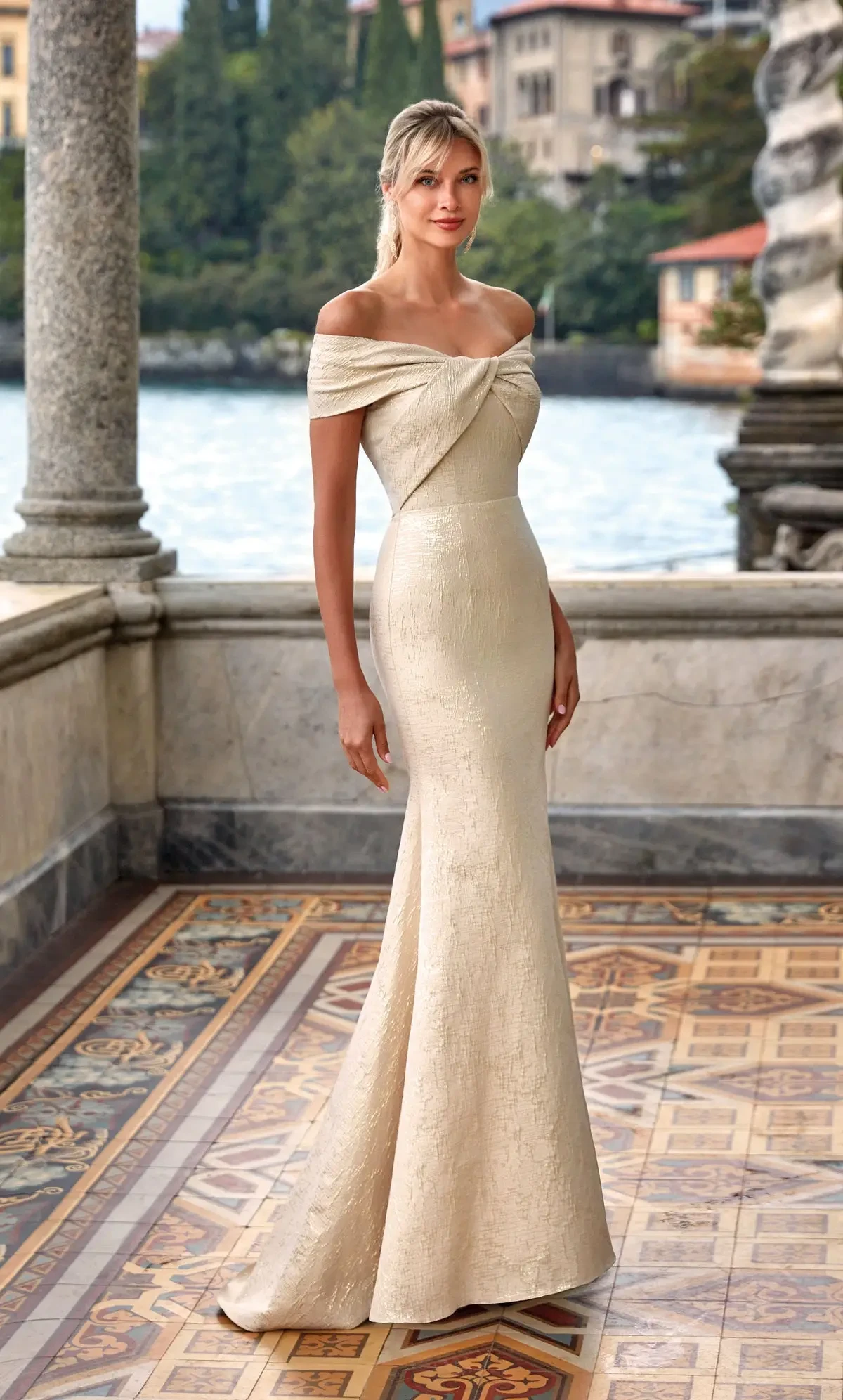 A woman in an elegant, off-the-shoulder gown stands on a balcony overlooking a body of water, surrounded by greenery and architecture. The dress is fitted and has a subtle sheen.