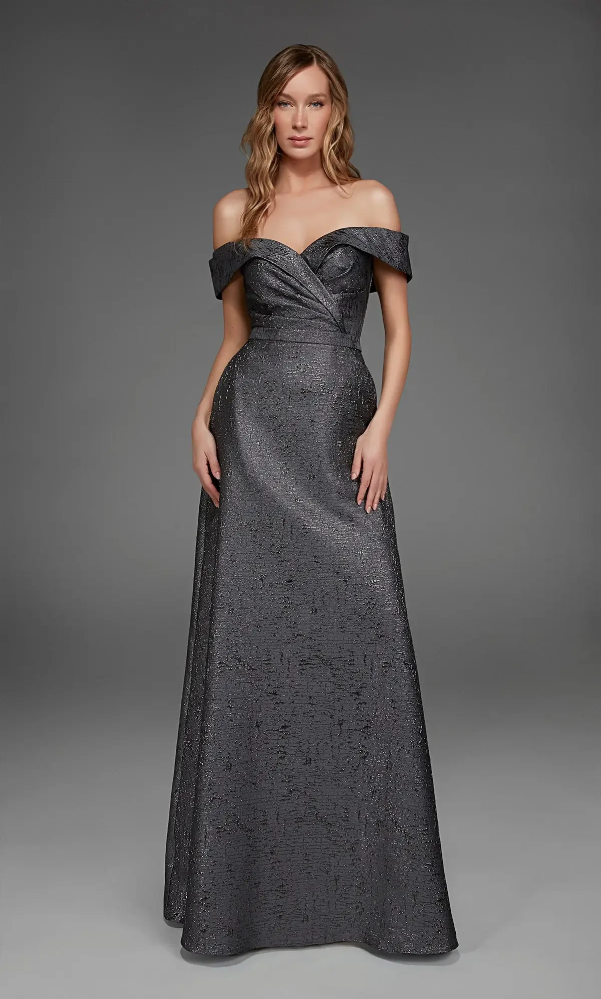 A woman wearing an elegant off-the-shoulder dark gray gown, standing against a neutral background. The dress features a fitted bodice and flows into a long skirt.