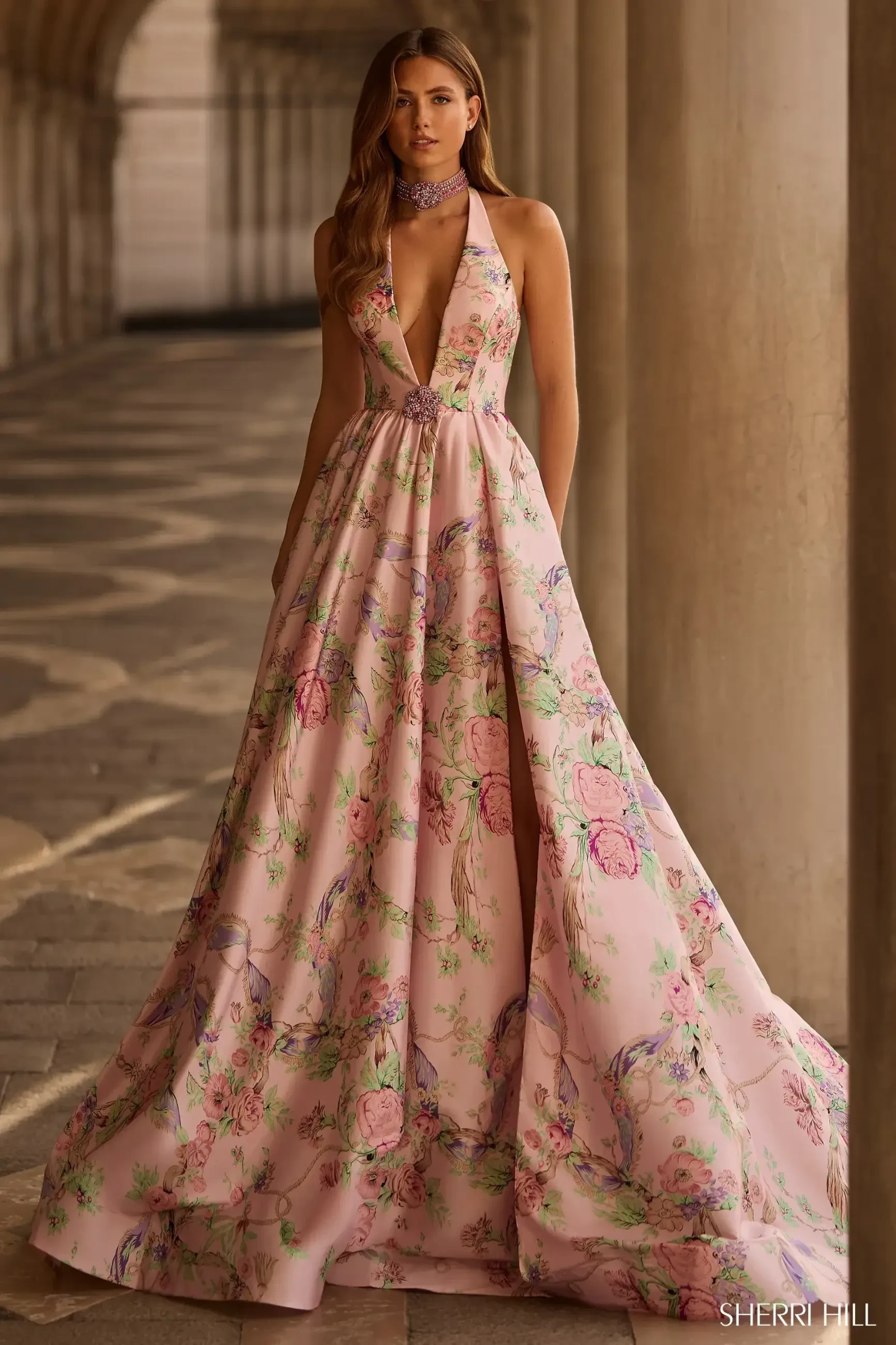 Woman in floral pink gown stands in an elegant, arched hallway. The dress features a deep V-neck and flowing skirt, creating a graceful, serene atmosphere.