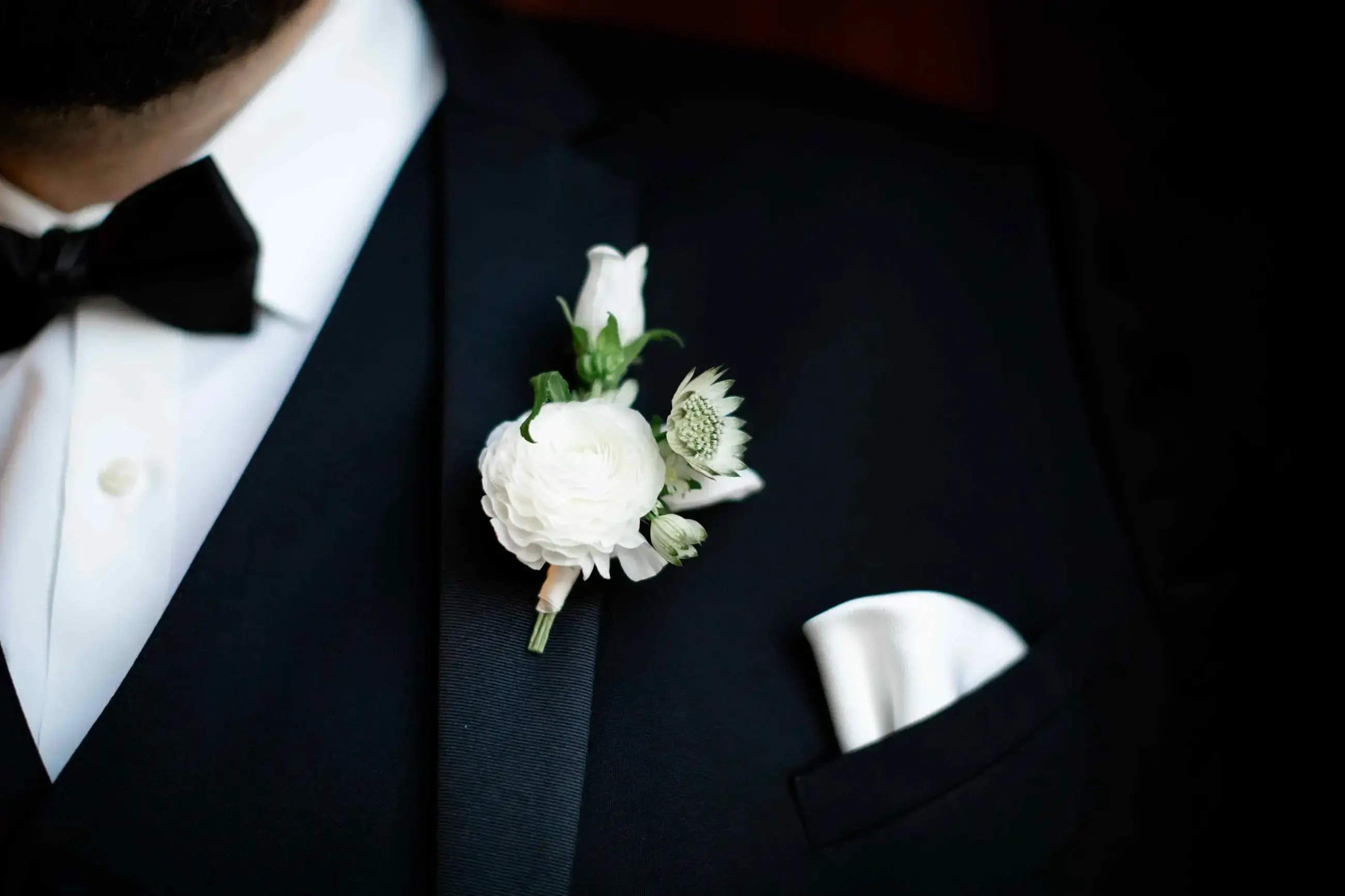 A close-up of a man's tuxedo, featuring a black bow tie and a white pocket square. There is a white flower boutonni&egrave;re pinned on the lapel.