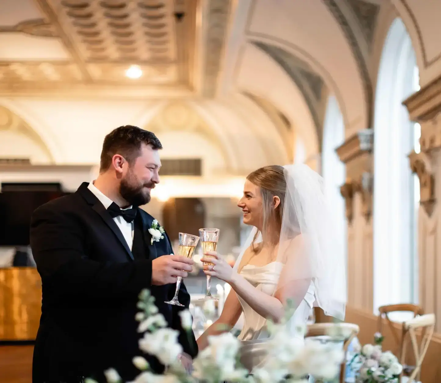 A bride and groom in formal attire clinking champagne glasses in a beautifully decorated venue. The bride is wearing a wedding dress and a veil, while the groom is dressed in a tuxedo. They are smiling at each other, celebrating their special moment.