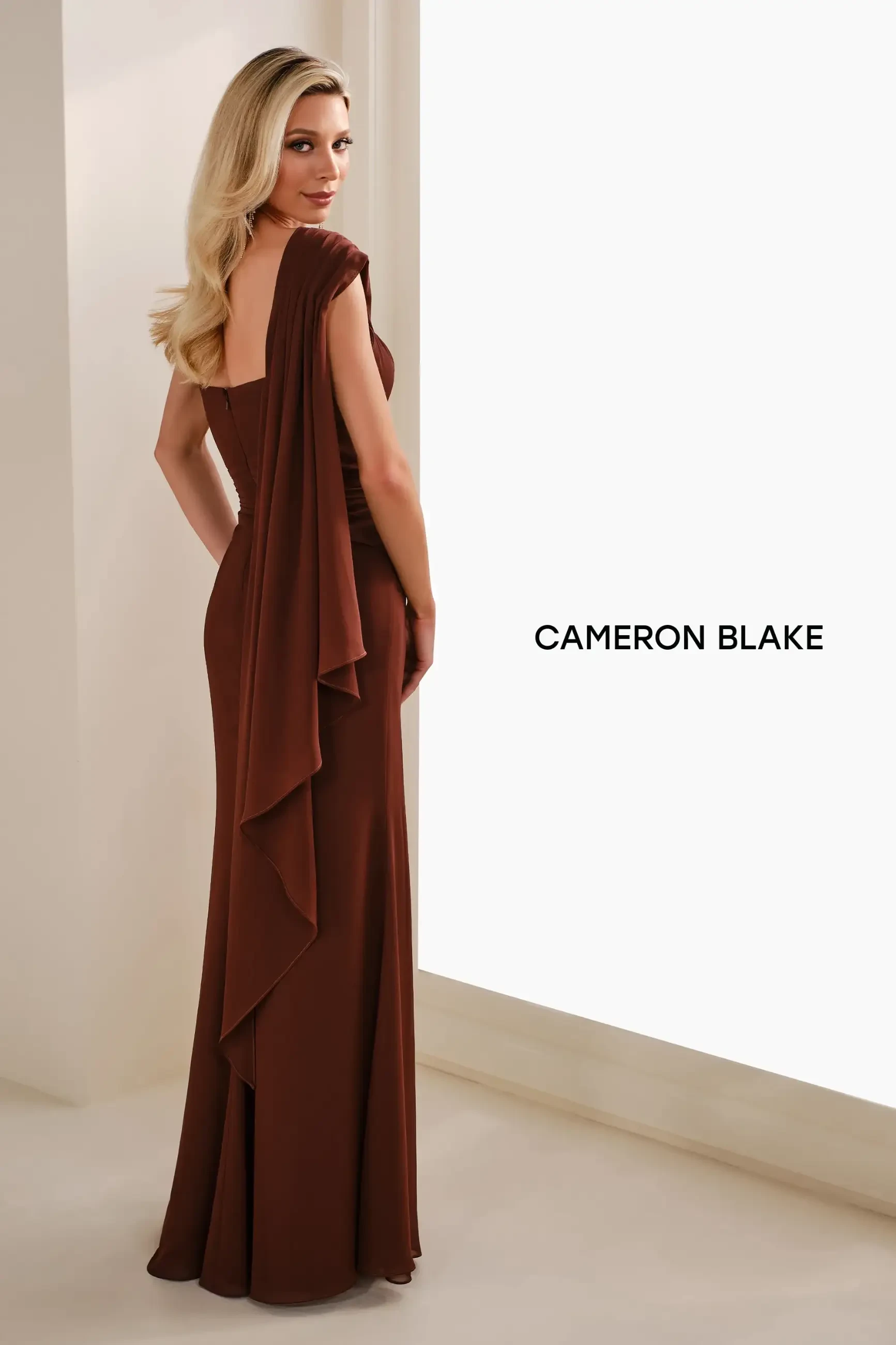 Model is wearing a cocoa chiffon sheath with a one-shoulder cape and delicate floral detail.