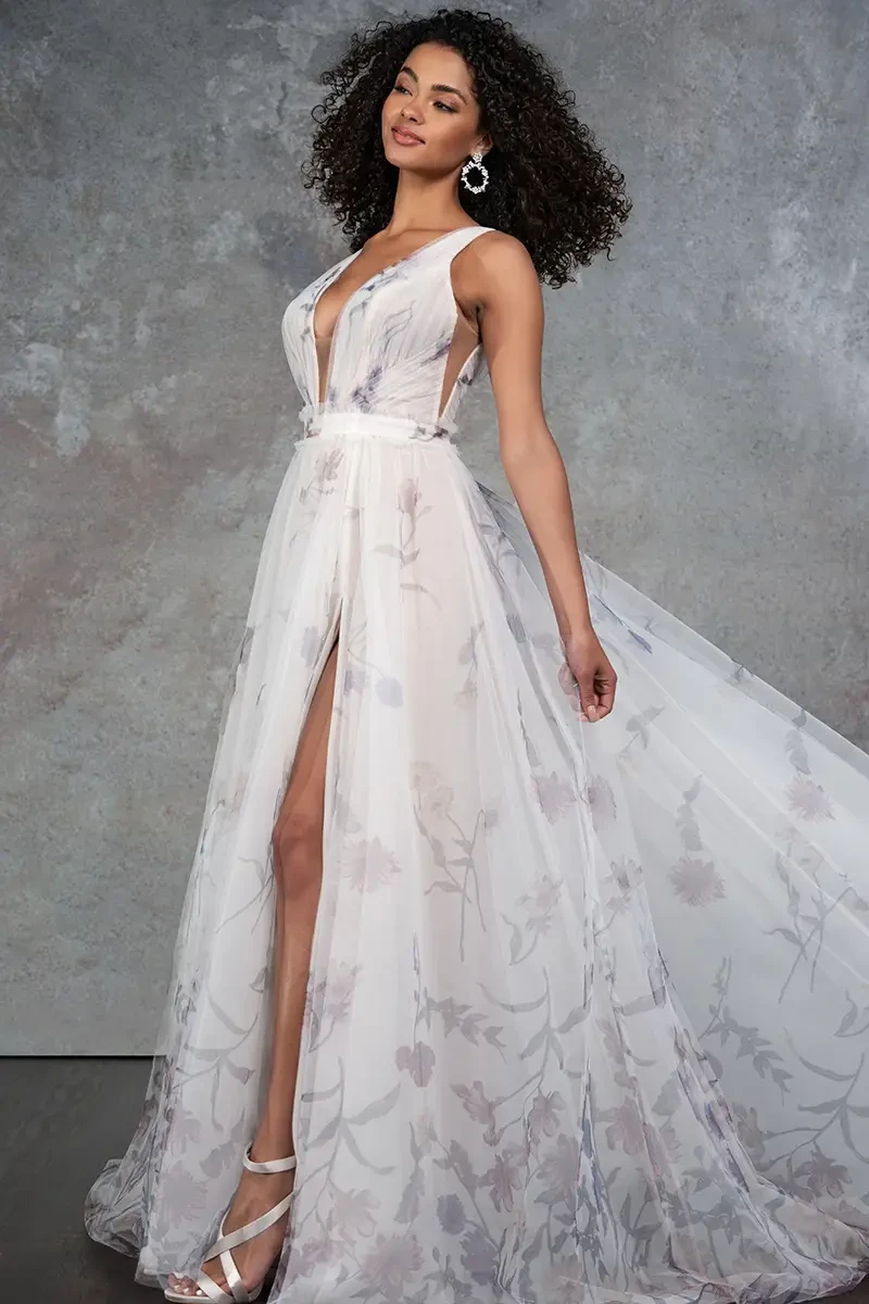 A woman poses confidently in a flowing, sheer floral gown with a high slit, against a textured gray backdrop. She wears statement earrings and heels.