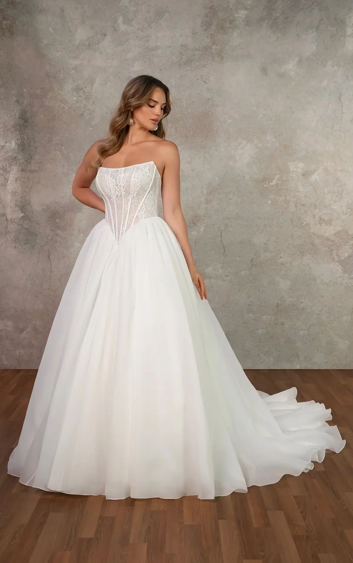 A bride wearing a strapless, lace bodice wedding gown with a flowing, full skirt stands on a wooden floor. The tone is elegant and serene.
