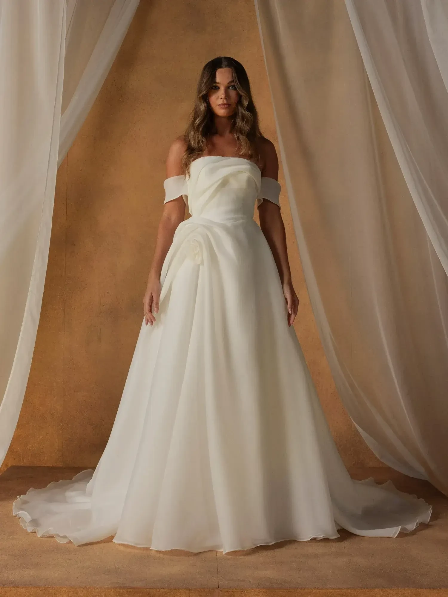 A woman in a strapless, off-the-shoulder wedding dress stands in front of a backdrop with draped fabric. The gown is flowing and elegant, and she appears confident and graceful.