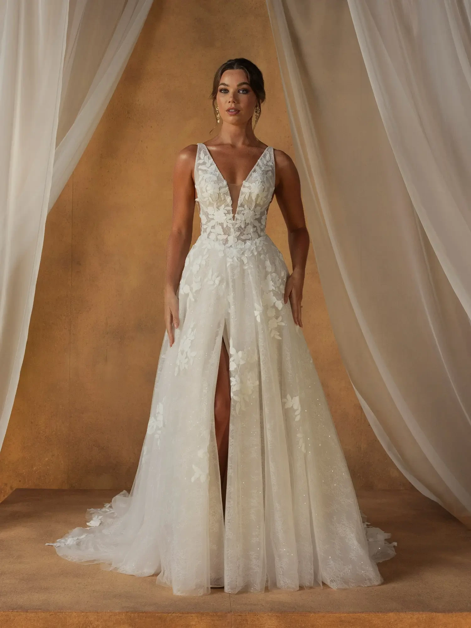 A woman stands confidently in a white floral wedding gown with a deep V-neck and high slit. The elegant backdrop features soft, flowing curtains.