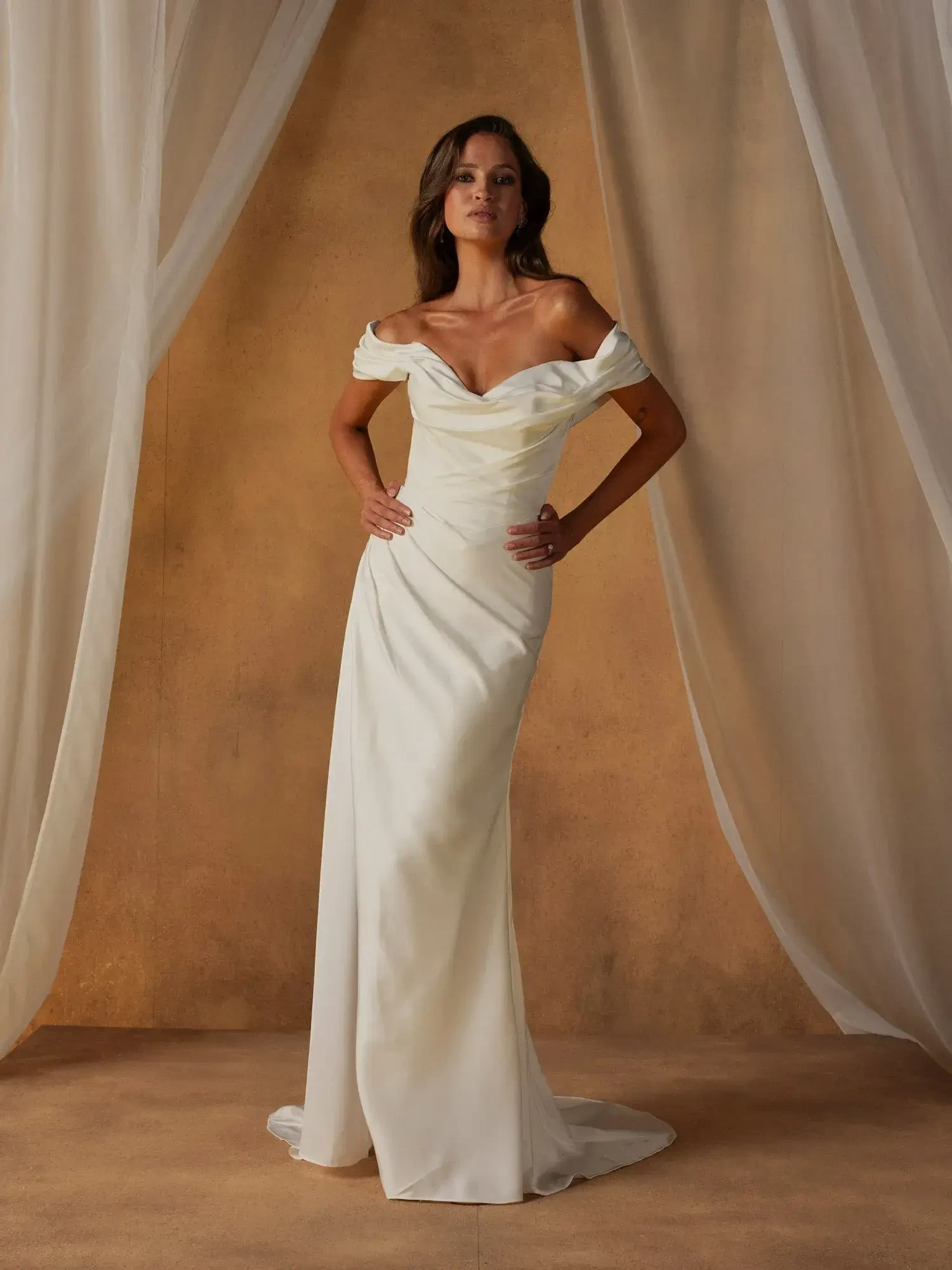 A woman stands in an elegant off-the-shoulder white dress, posing confidently against a warm-toned background with sheer drapery.