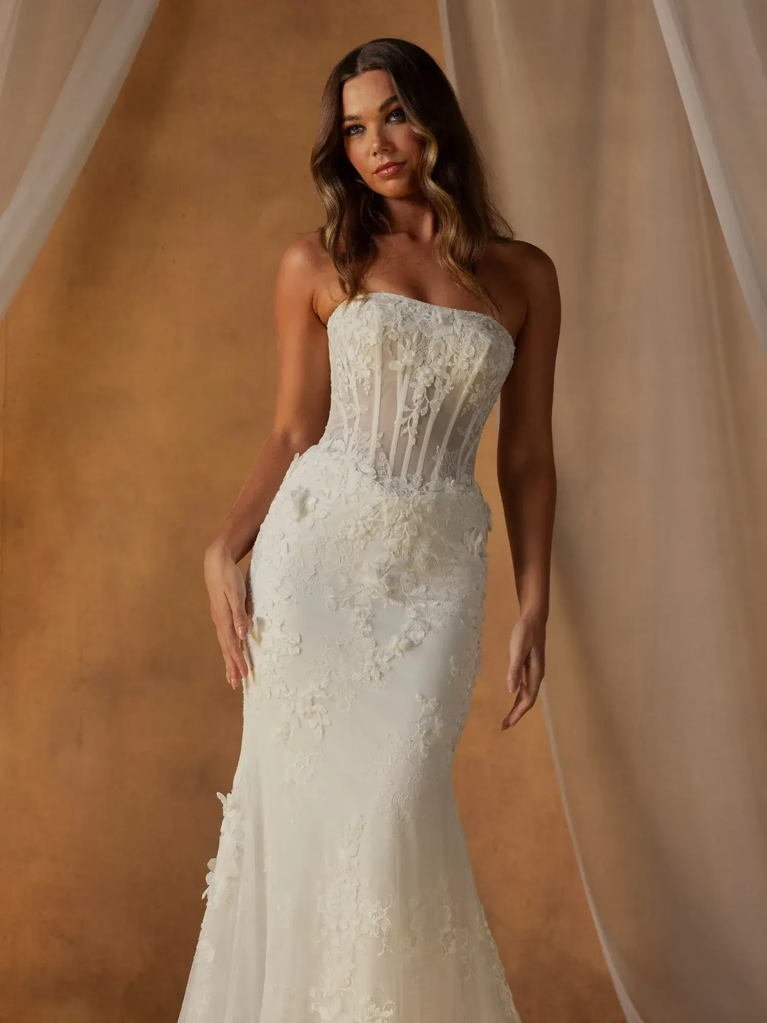 A woman wearing a strapless white gown with intricate lace and floral details, standing elegantly against a softly lit backdrop.